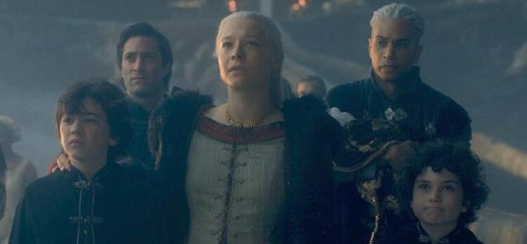 Rhaenyra on the stone bridge to Dragonstone, looking upward, with arms around two of her sons and Laenor slightly behind her with the baby. An entourage of others are behind them.