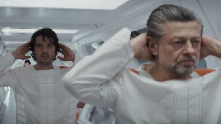 Cassian walks behind his floor manager in a prison corridor. Both of them have their hands behind their heads.