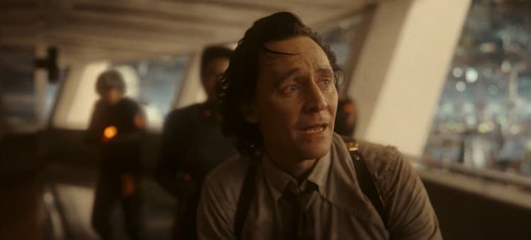 Loki is looking to his left and has a concerned look on his face. TVA Agents can be seen behind him and they are in a hallway with a wall of windows.