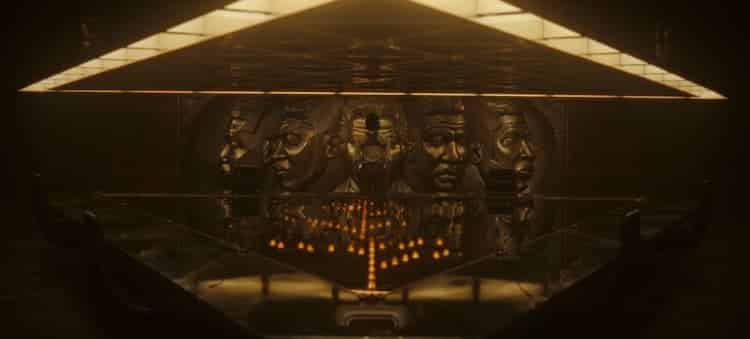 Loki is in a conference room that has a wall decorated with Kang's face. A triangle desk is in the centre of the room.