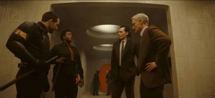 Hunter B-15, Loki, Mobius, and another Hunter are standing outside a holding cell in the TVA. An orange door is down the hall and some light is coming in from above.