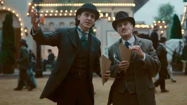 Loki and Mobius are eating snacks. A building covered with lights is behind them. People are walking in both directions behind them as well.