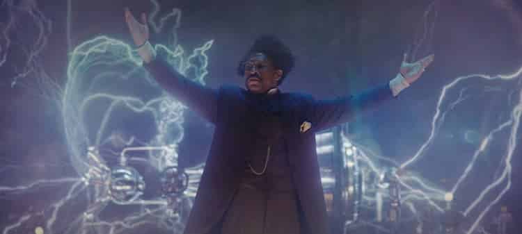 Victor Timely is on a stage giving a presentation. His invention is behind him and the heads of some people can be seen at the bottom of the image.