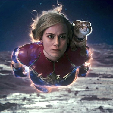 Carol is covered in orange and blue energy. She is flying across the surface of a planet with a concerned look on her face. Goose the cat is sitting on her left shoulder with their eyes closed.