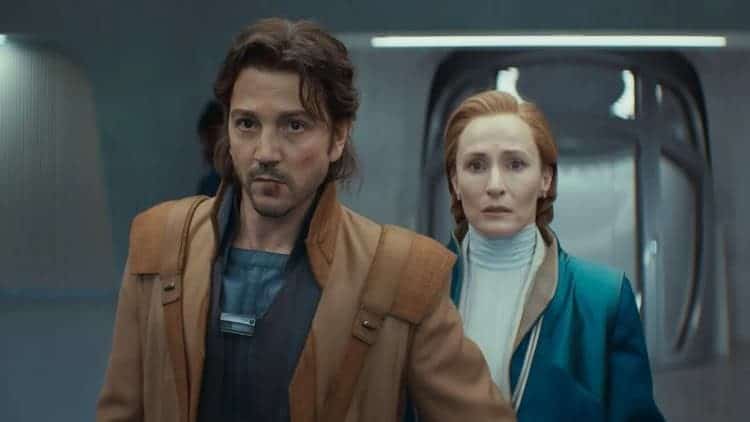 Cassian and Mon Mothma are in the lobby of the Senate building. Cassian has a focused look on his face while Mothma looks concerned. Cassian has injuries on his face. The area is dimly lit.
