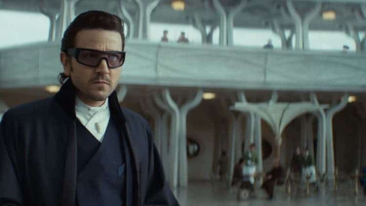 Cassian is in disguise on Ghorman. He is wearing a fashionable jacket with a fancy pair of sunglasses. He is walking through the plaza in the middle of the day. He has a serious look on his face and is looking around the area.