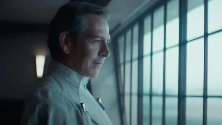 Director Krennic is looking out a window. It's a cloudy day, and not much light is entering the room. He has a smile on his face.