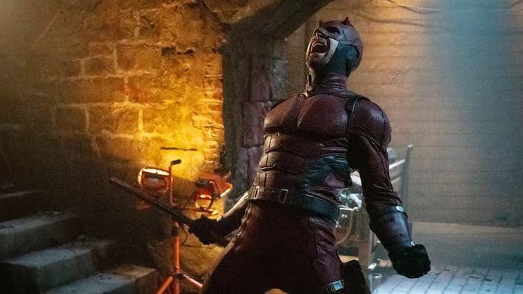 Daredevil is screaming in an underground lair. He's holding one of his batons in his right hand. Work lights are illuminating the area. Brick walls line the surroundings.