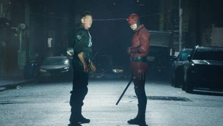 The Punisher and Daredevil are standing in the middle of the street at night. They are looking at the same thing coming towards them. They both look injured. Daredevil is holding his weapon in his right hand.