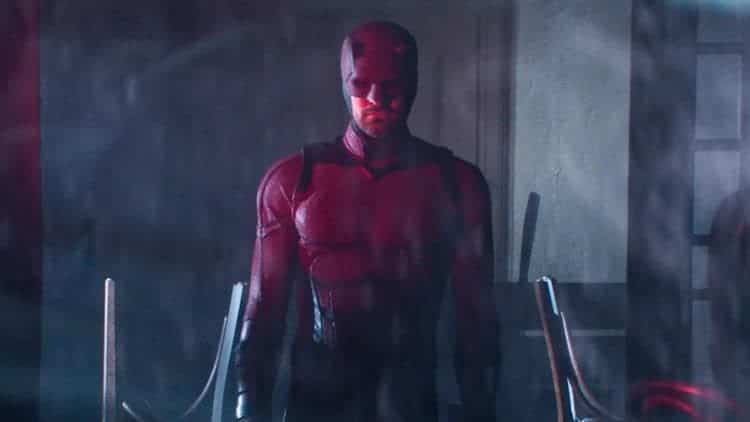Daredevil is standing in a closed bar. The chairs behind him are upside down on the table. A red light from outside illuminates parts of his body.